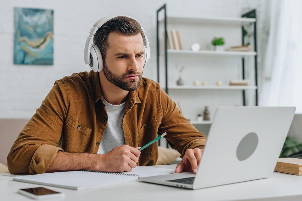 Mann lernt am Laptop mit Kopfh&ouml;rern die Unterschiede zwischen Bayerisch und Hochdeutsch und macht sich Notizen.